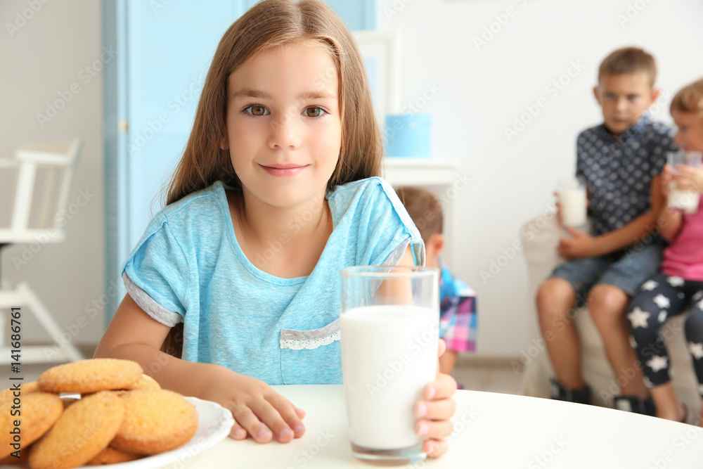 Cute little girl with glass of milk sitting at table in the room