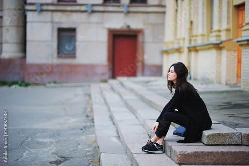 Portrait of young beautiful dark-haired girl with a sad look, sitting on the steps in courtyard buildings, side view