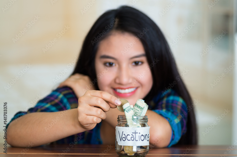 Young brunette woman sitting down facing camera, putting coins inside glass jar with money inside, label reading vacations, smiling happily Young brunette woman sitting down facing camera, putting coins inside glass jar with money inside, label reading vacations, smiling happily