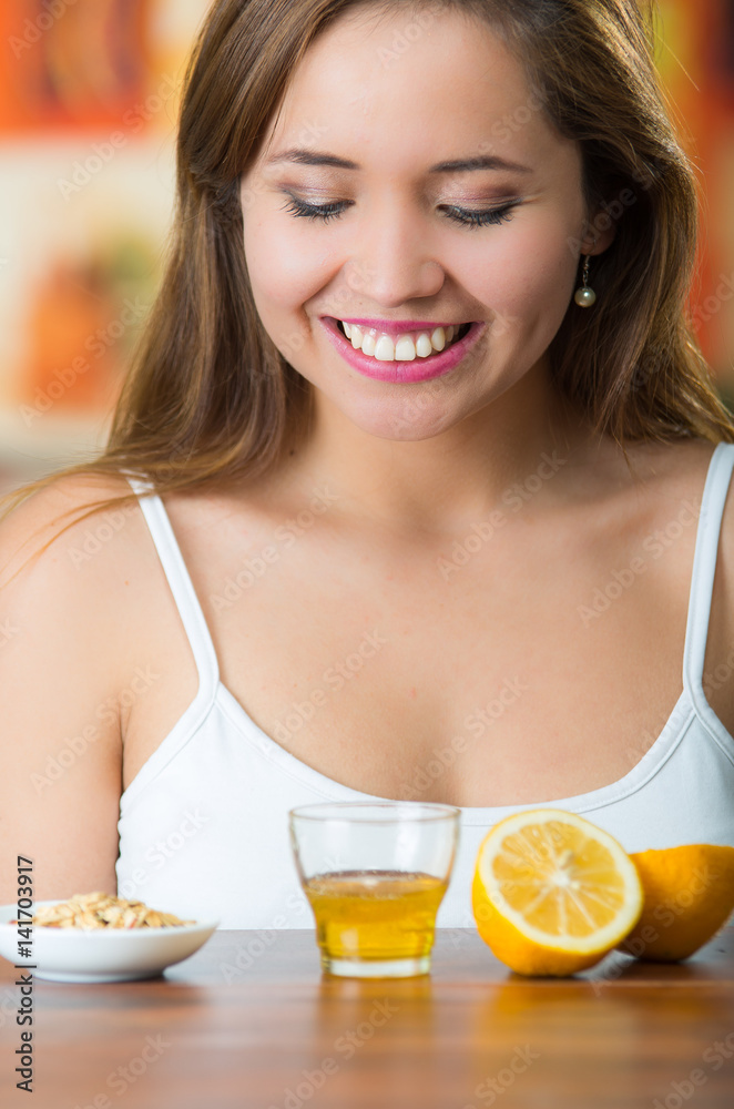 Young brunette sitting by table with glass of honey, granola and sliced lemon in front, smiling happily Young brunette sitting by table with glass of honey, granola and sliced lemon in front, smiling happily