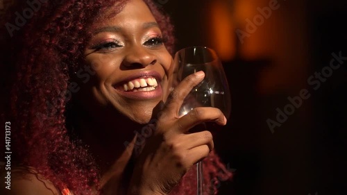 The charming smile of the young afro-american girl with the red curly hair holding the wineglass with red wine in the cafe. Close-up portrait photo