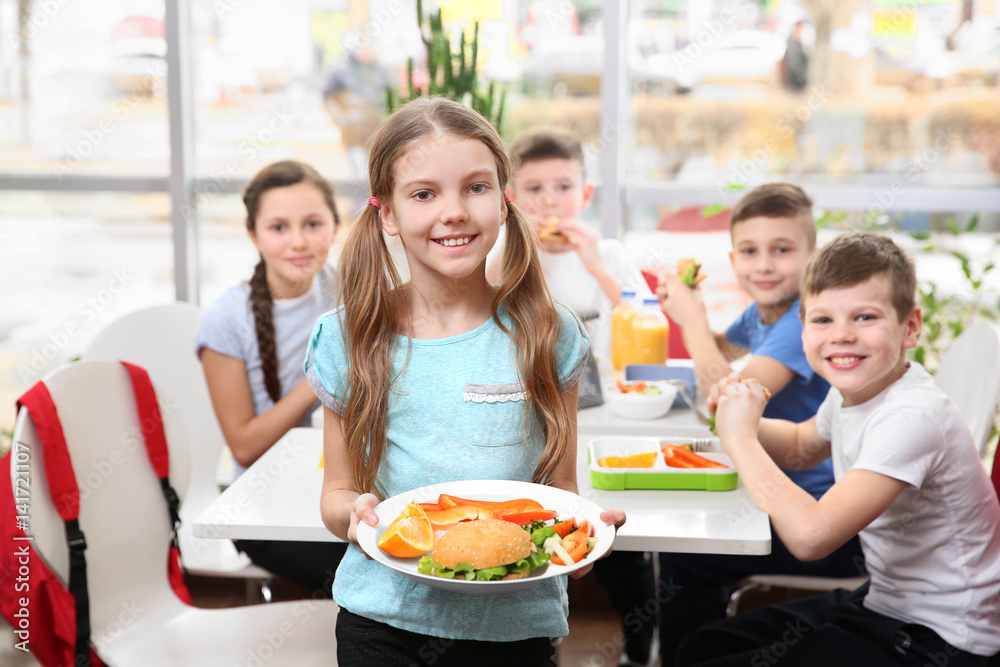 Cute girl holding plate with delicious food and children eating at table in school cafeteria