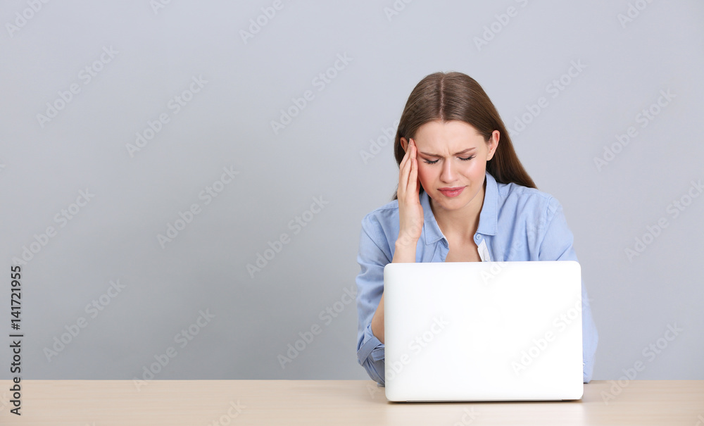 Beautiful young woman suffering from headache while working with laptop, on grey background Beautiful young woman suffering from headache while working with laptop, on grey background