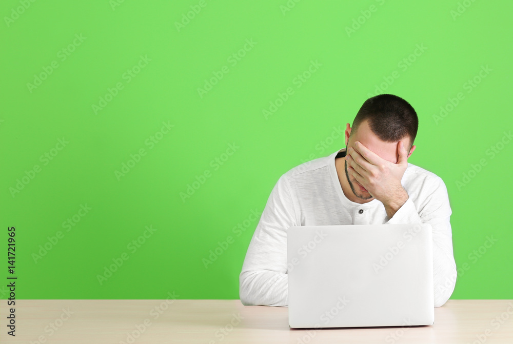 Handsome young man suffering from headache while working with laptop, on color background