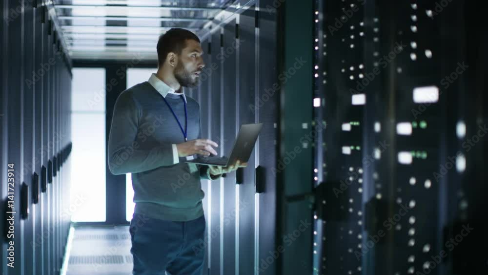Male Server Engineer in Data Center. He Stands Before Open Door of Rack Server and Works on His Laptop. Shot on RED EPIC-W 8K Helium Cinema Camera.