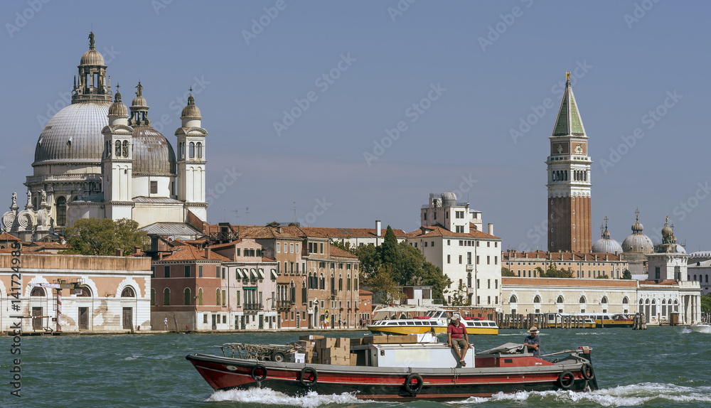 A boat with a man sitting on it crosses the Venetian lagoon with in the background the Campanile di San Marco and the Basilica della Salute, Venice, Italy