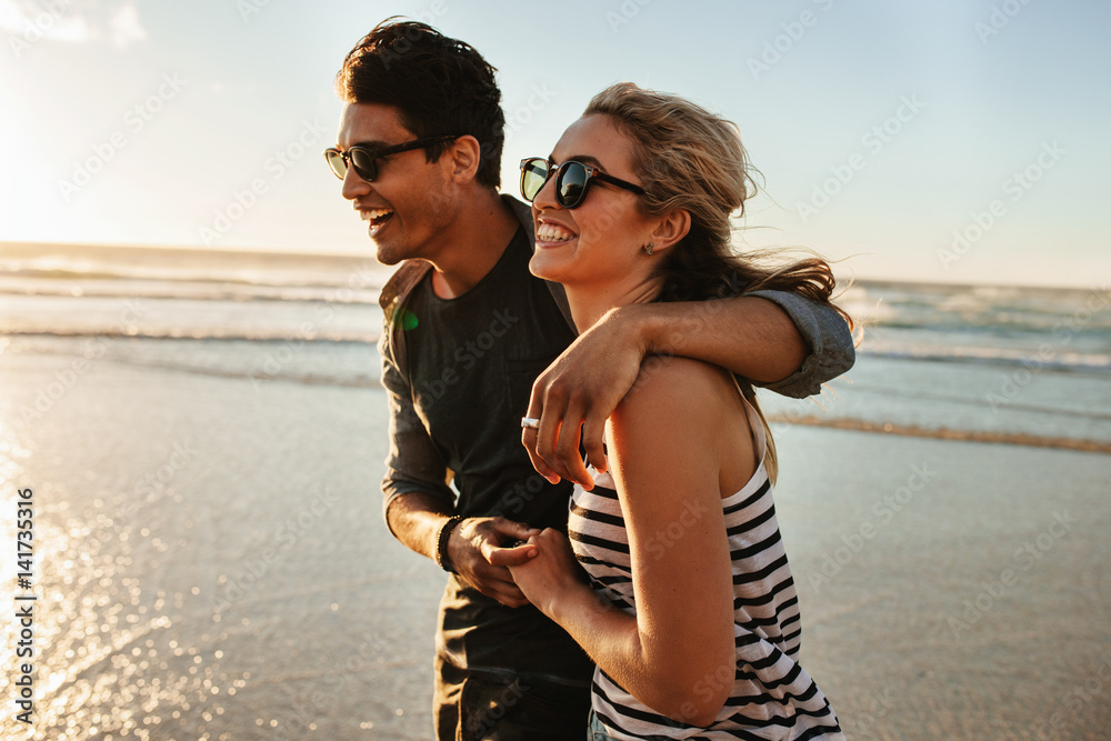 Smiling young couple walking on beach - obrazy, fototapety, plakaty 