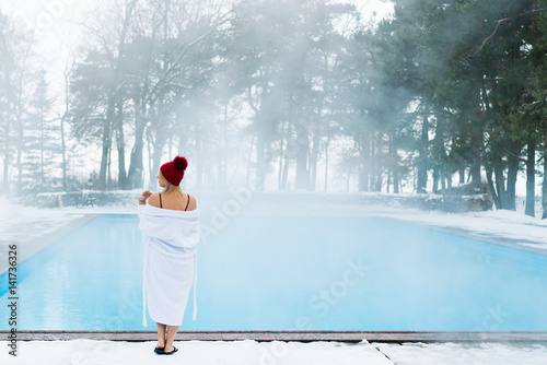 Young blonde woman in bathrobe and red hut near outdoor swimming pool at winter day