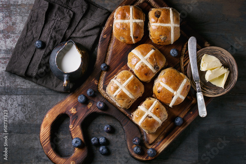 Hot cross buns on wooden cutting board served with butter, knife, fresh blueberries and jug of cream on textile papkin over old texture metal background. Top view, space. Easter baking.