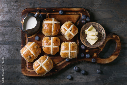 Hot cross buns on wooden cutting board served with butter, knife, fresh blueberries and jug of cream over old texture metal background. Top view, space. Easter baking.