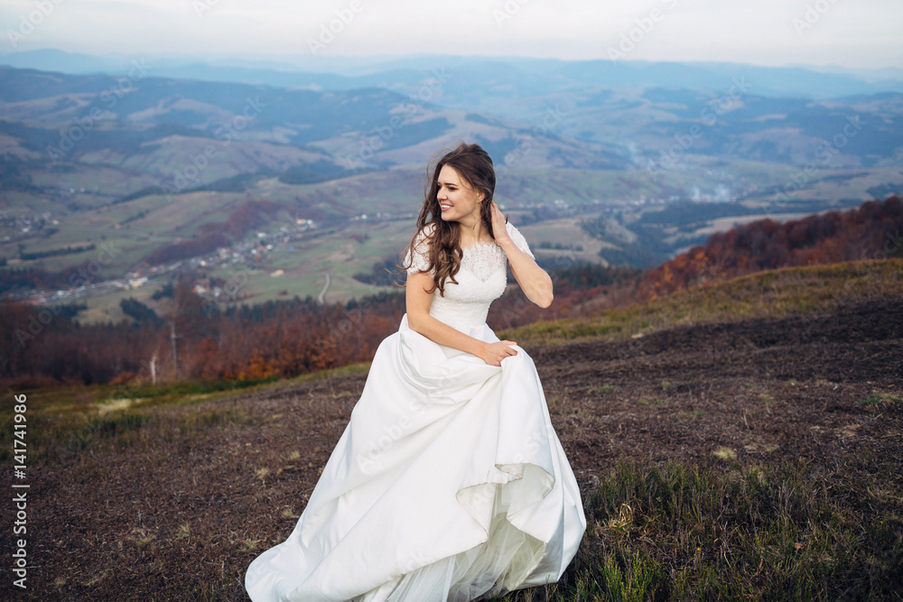 Joyful bride looks over her shoulder posing on autumn hill with great mountain view behind her