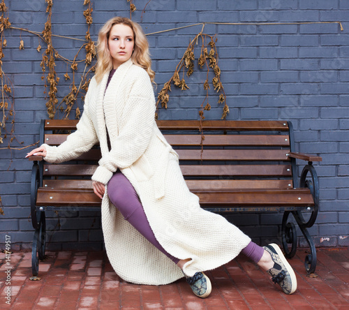 Beautiful blonde girl posing sitting on a bench in a fashionable knitted long coat anf oufit near a gray brick wall. Fashion Style photo