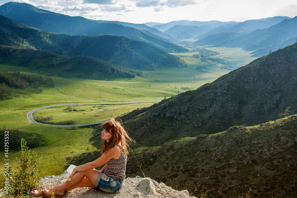The girl at the cliff on the mountain. Tourist in shorts sitting near the edge of the cliff. At the bottom of a beautiful valley with green hills and mountains and winding road. Mountain Altai, Russia