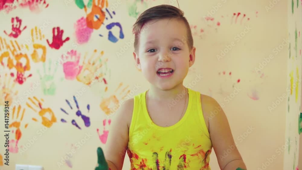 Close up veiw of cute little boy with painted hands after leaving his colorful handprints on the wall and showing his thumb up. Young happy family. Mother and child concept Close up veiw of cute little boy with painted hands after leaving his colorful handprints on the wall and showing his thumb up. Young happy family. Mother and child concept