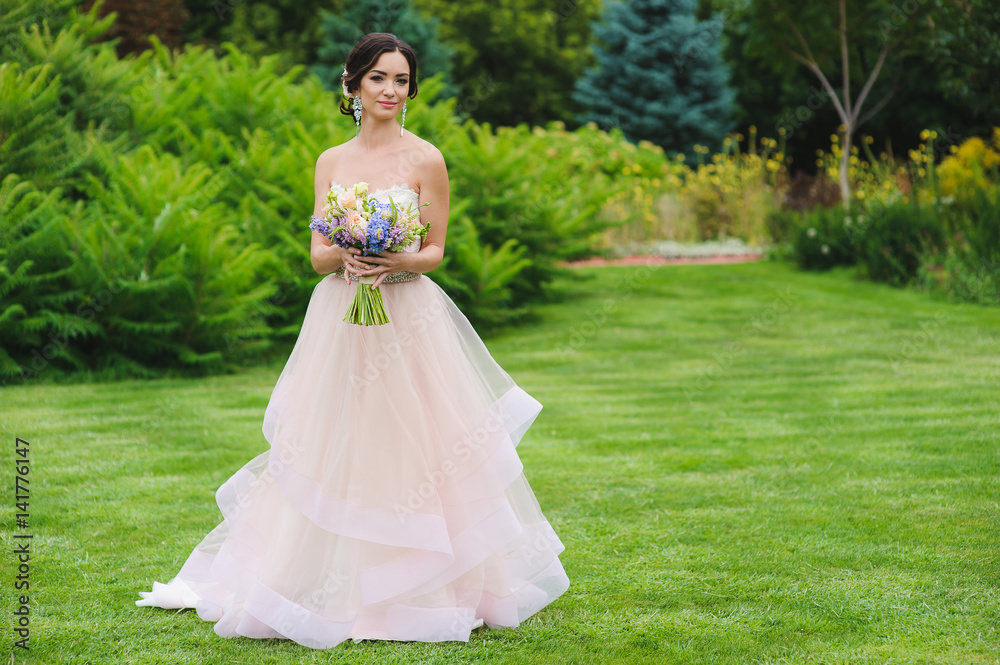 Portrait of a beautiful bride in park. Cute lady with a bouquet in pink wedding dress outdoors. Looking sideways. Grass, trees and bushes in the background.