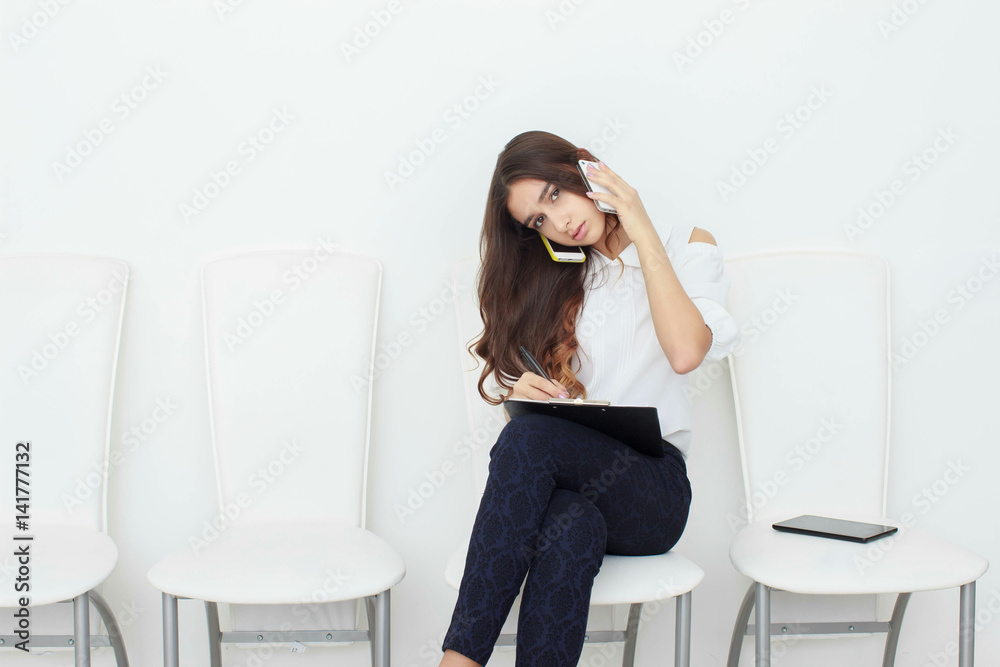 The girl is sitting on a white chair talking on the phone and writing in the tablet. Multitasking The girl is sitting on a white chair talking on the phone and writing in the tablet. Multitasking