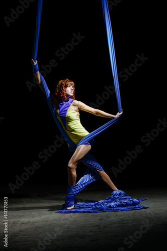 Beautiful young athletic girl, with redhead in yellow costume, posing with gorgeous blue aerial silks on the light in the darkness. Diagonal with canvases wrapped around slender legs.