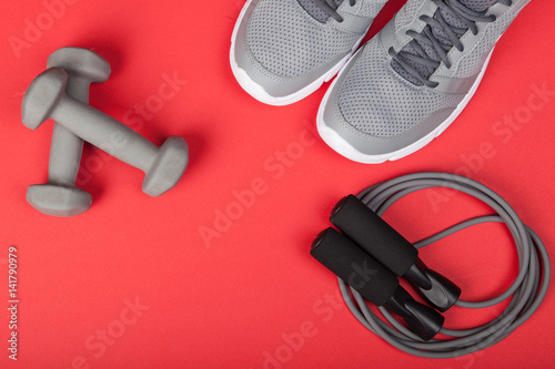 Sport shoes, dumbbells and skipping rope on red background. Top view. Fitness, sport and healthy lifestyle concept.