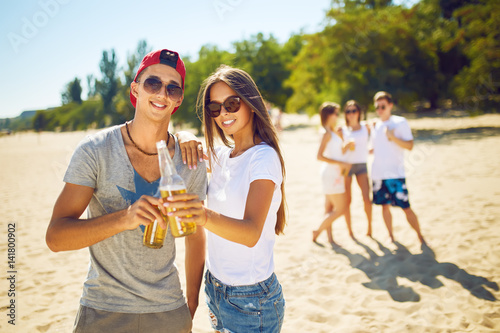 boy and a girl are holding bottles of beer in hands and smiling .Group of friends hanging out with beer at the beach.  Excellent sunny weather. Beautiful figures. Super mood. Summer concept   © maxbelchenko