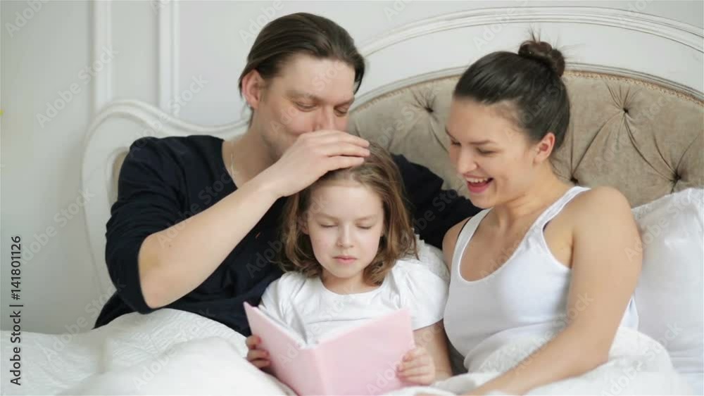 Beautiful Little Girl is Reading Aloud the Book Lying on the Bed with Her Parents. Handsome Father is Kissing His Clever Child. Beautiful Little Girl is Reading Aloud the Book Lying on the Bed with Her Parents. Handsome Father is Kissing His Clever Child.