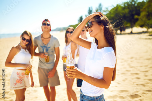Beautiful girl with glasses is holding a bottle with beer in her hands. Group of friends hanging out with beer at the beach. Excellent sunny weather. Beautiful figures. Super mood. Summer concept