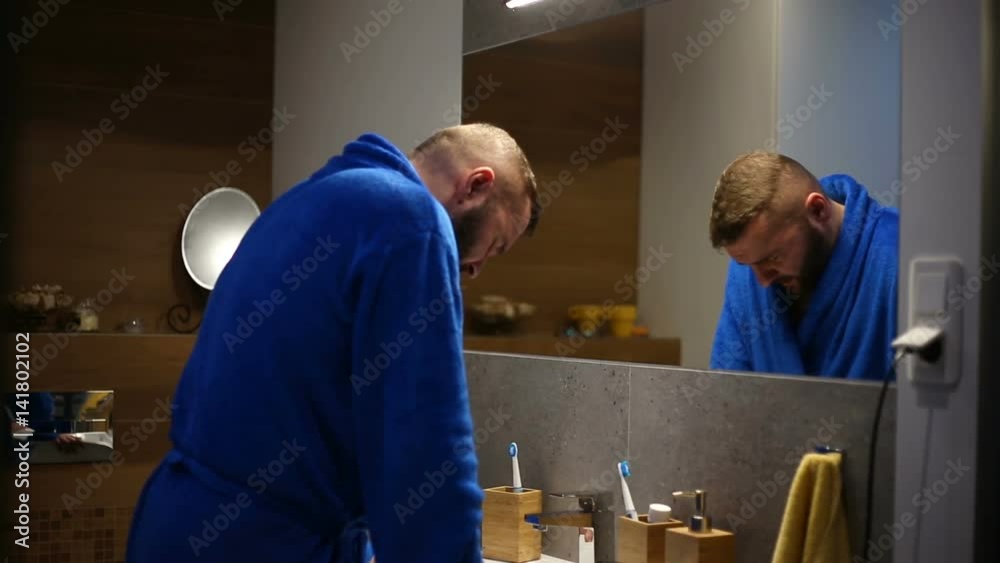 Worried man wearing bathrobe and having some problems while looking to the mirror

