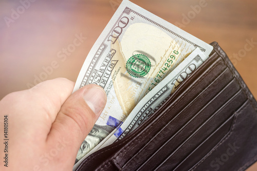 Close-up of male hand taking hundred dollar banknotes from wallet, wooden background, money and finance concept.