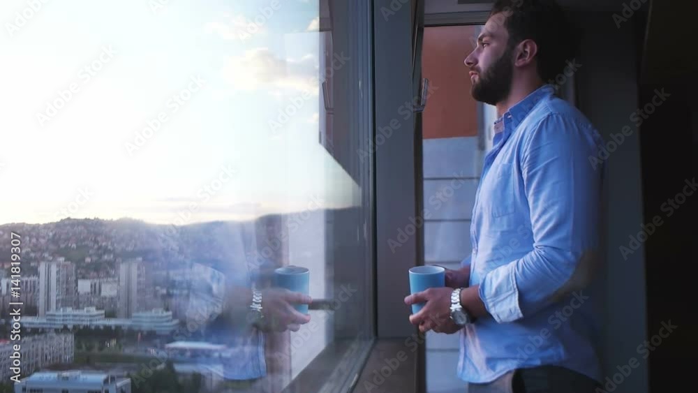 Man in shirt drinking coffee or tea by window admiring greath view on town during sunset Man in shirt drinking coffee or tea by window admiring greath view on town during sunset