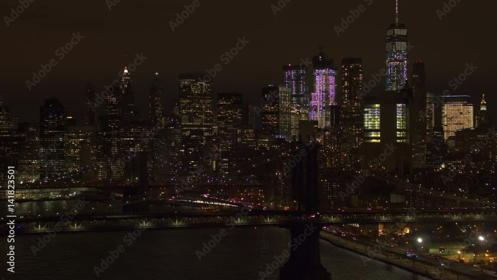 AERIAL HELI SHOT: Automobile traffic moving across the iconic Brooklyn and Manhattan bridge lit up with lights on magical summer night in New York City overlooking lighted Downtown financial district AERIAL HELI SHOT: Automobile traffic moving across the iconic Brooklyn and Manhattan bridge lit up with lights on magical summer night in New York City overlooking lighted Downtown financial district