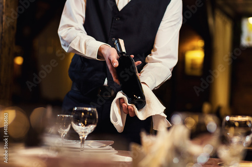 The waiter offers visitors wine,Waiter in uniform waiting an order,Waiter with a white towel on his hand,Confident waiter,A pub.Restaurant.Classic.Evening.European restaurant photo