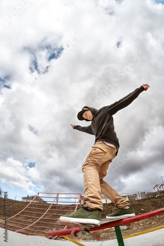 Teen skater in a hoodie sweatshirt and jeans slides over a railing on a skateboard in a skate park