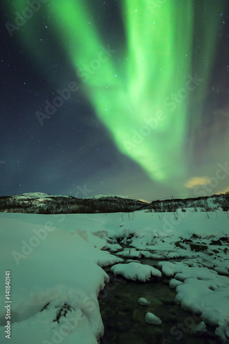 Northern lights Aurora Borealis during night above a water stream in a winter landscape.