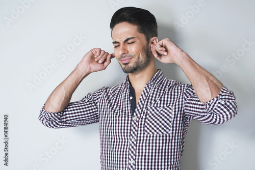 Stop that sound! Close up portrait of young man, holding hands to ears covering to stop noise, looking stressed in unbutton shirt against grey background. Human emotions extremes