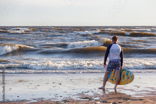 Young sportive man is running after his yellow and blue skimboard on the beach on a sunny day and on the sky there are dark clouds