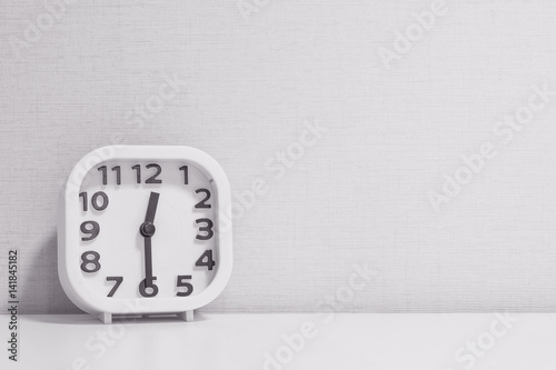 Closeup white clock for decorate show half past twelve or 12:30 p.m. on white wood desk and cream wallpaper textured background in black and white tone with copy space