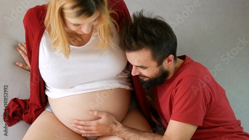 Happy bearded man touching pregnant woman belly, young excited couple expecting baby relaxing together on the bed at home.
