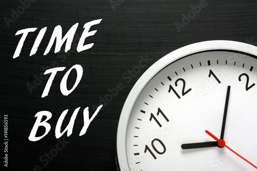 The phrase Time to Buy written on a blackboard next to a modern wall clock as a reminder for when to invest