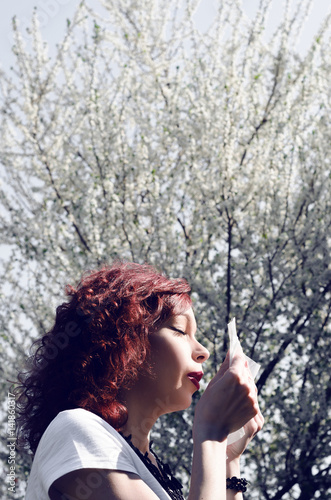 beautiful woman with red hair standing under a tree flower in spring and blowing nose, hayfever