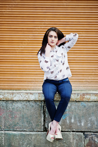 Young stylish brunette girl on shirt, pants and high heels shoes, sitting and posed background orange shutter. Street fashion model concept.