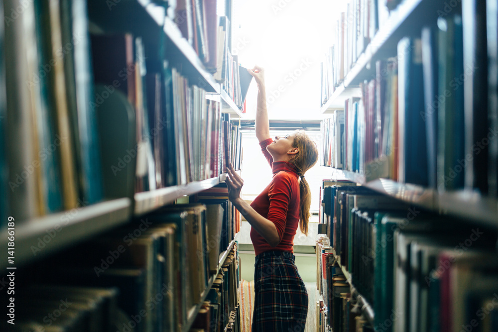 Young librarian searching books and taking one book from library bookshelf - obrazy, fototapety, plakaty 