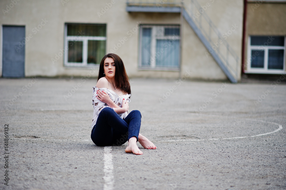 Young stylish teenage brunette girl on shirt, pants and high heels shoes, sitting on pavement and posed background school backyard. Street fashion model concept. Young stylish teenage brunette girl on shirt, pants and high heels shoes, sitting on pavement and posed background school backyard. Street fashion model concept.