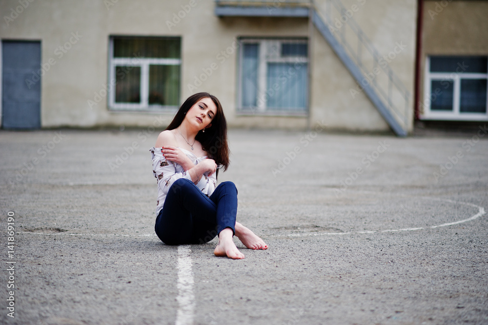 Young stylish teenage brunette girl on shirt, pants and high heels shoes, sitting on pavement and posed background school backyard. Street fashion model concept.