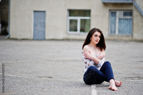 Young stylish teenage brunette girl on shirt, pants and high heels shoes, sitting on pavement and posed background school backyard. Street fashion model concept. © AS Photo Family