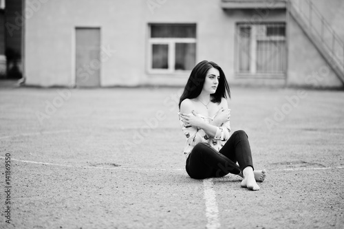 Young stylish teenage brunette girl on shirt, pants and high heels shoes, sitting on pavement and posed background school backyard. Street fashion model concept.