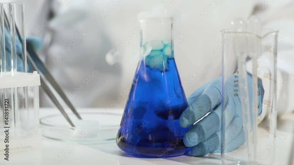 Lab assistant putting dry ice into blue liquid in conical flask, making it boil Lab assistant putting dry ice into blue liquid in conical flask, making it boil