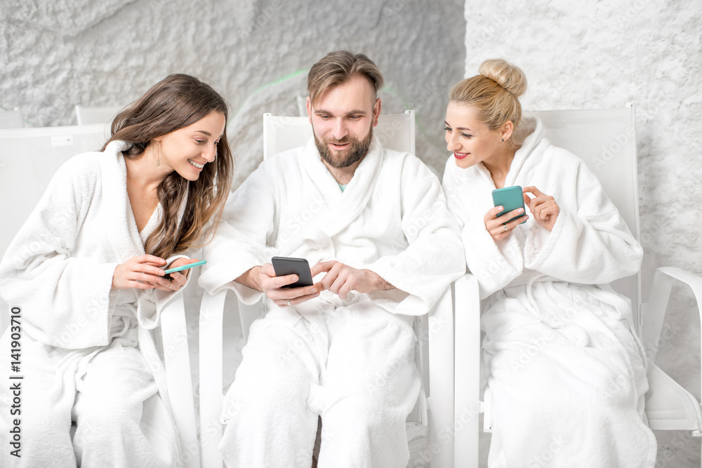 Three friends in bathrobes sitting with phones, applying salt therapy in the salt room Three friends in bathrobes sitting with phones, applying salt therapy in the salt room