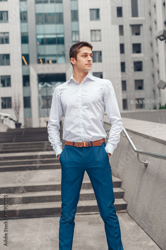 Portrait of cheerful businessman standing outdoors near the business center. Young man holds hands in his trouser pockets.