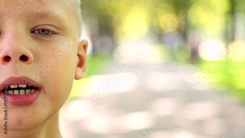 Closeup portrait of happy smiling cute boy with short blond hair. Caucasian child posing for camera in city park on beautiful sunny summer day. Real time full hd video footage with copy space.