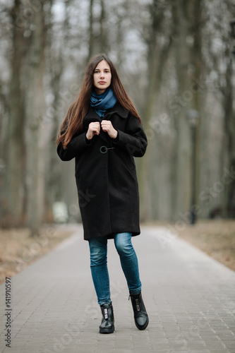 Young beautiful girl in a black coat and blue scarf for a walk in the autumn / spring park. An elegant brunette girl with gorgeous extra long hair. Lifestyle concept.