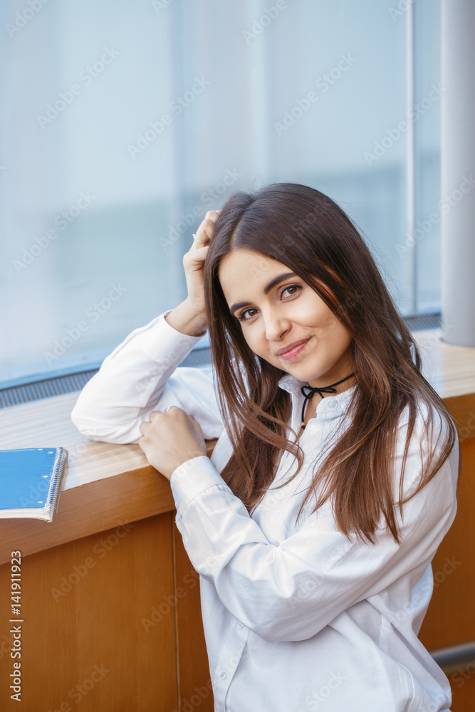 Portrait of smiling Caucasian brunette young beautiful girl woman student with long dark hair and brown eyes in white shirt in college university looking in camera Portrait of smiling Caucasian brunette young beautiful girl woman student with long dark hair and brown eyes in white shirt in college university looking in camera